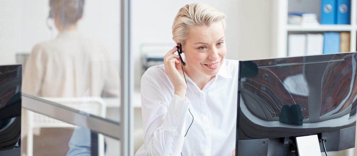 Portrait of smiling businesswoman speaking to microphone while using computer in office interior, customer support concept, copy space